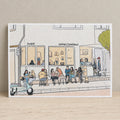An art print featuring a hand-drawn sketch of a street scene with a coffee shop, seated people, a scooter, and a tree, displayed on a light wooden surface with a blurred outdoor background.