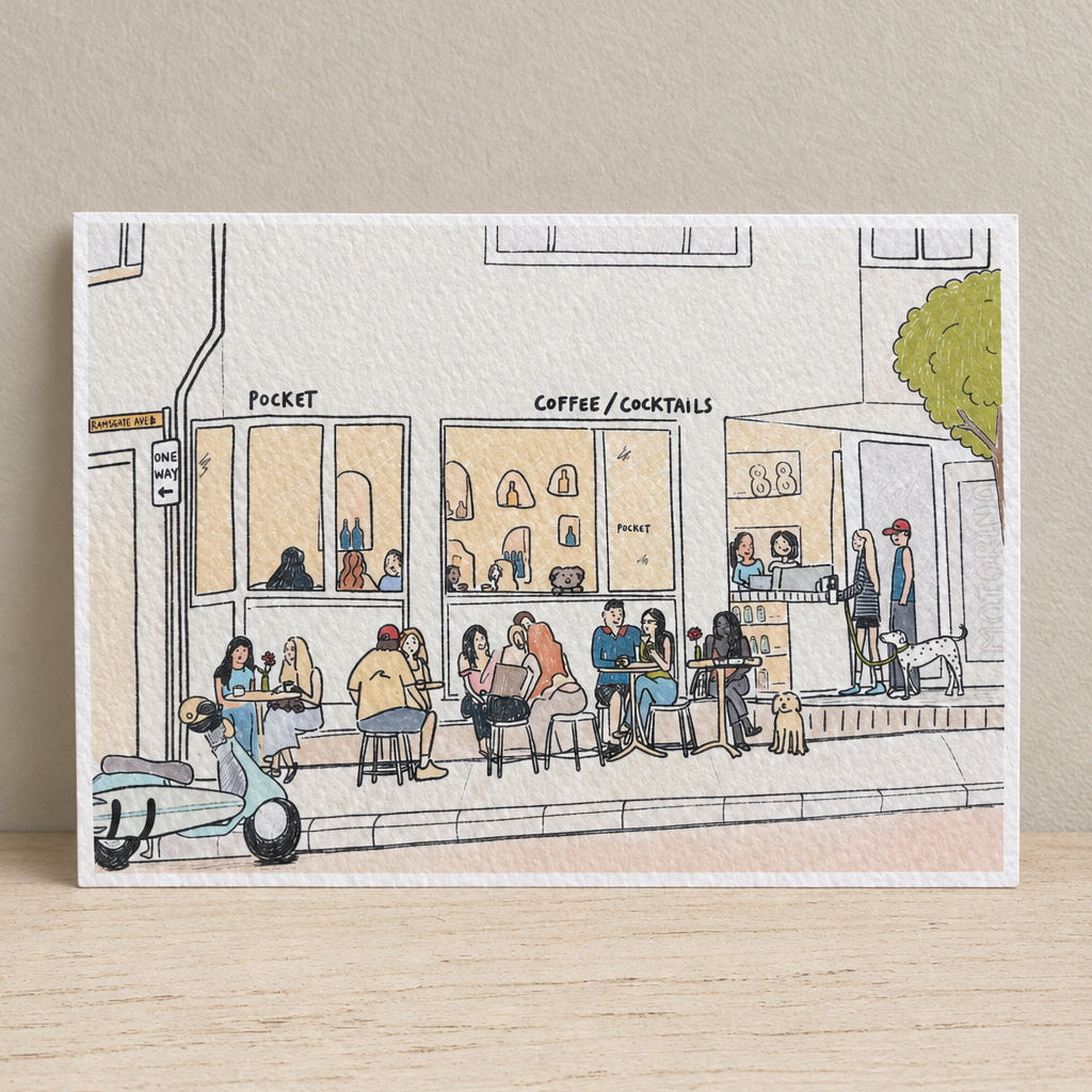 An art print featuring a hand-drawn sketch of a street scene with a coffee shop, seated people, a scooter, and a tree, displayed on a light wooden surface with a blurred outdoor background.